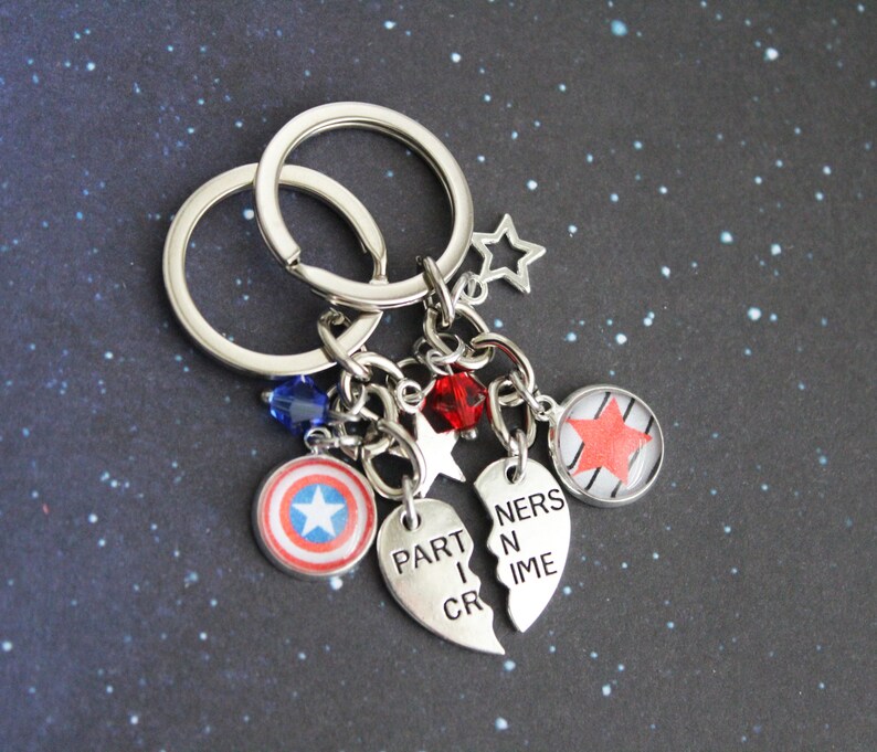 K&ouml;nnte beinhalten: Silberne Schl&uuml;sselanh&auml;nger mit Anh&auml;ngern. Zu den Anh&auml;ngern geh&ouml;ren ein Captain America-Schild, ein roter Stern und zwei halbherzf&ouml;rmige Anh&auml;nger mit der Aufschrift "PARTNERS IN CRIME". Zus&auml;tzliche Anh&auml;nger sind blaue und rote Perlen sowie sternf&ouml;rmige Anh&auml;nger.