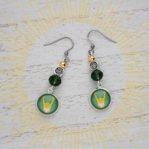 以下が含まれることがあります： 緑と金のアクセントが施されたシルバーカラーのドロップピアス。各イヤリングには、金のデザイン、緑色のビーズ、ケルトノットチャームが付いた円形の緑色のチャームが付いています。スタイリッシュなアクセサリー。