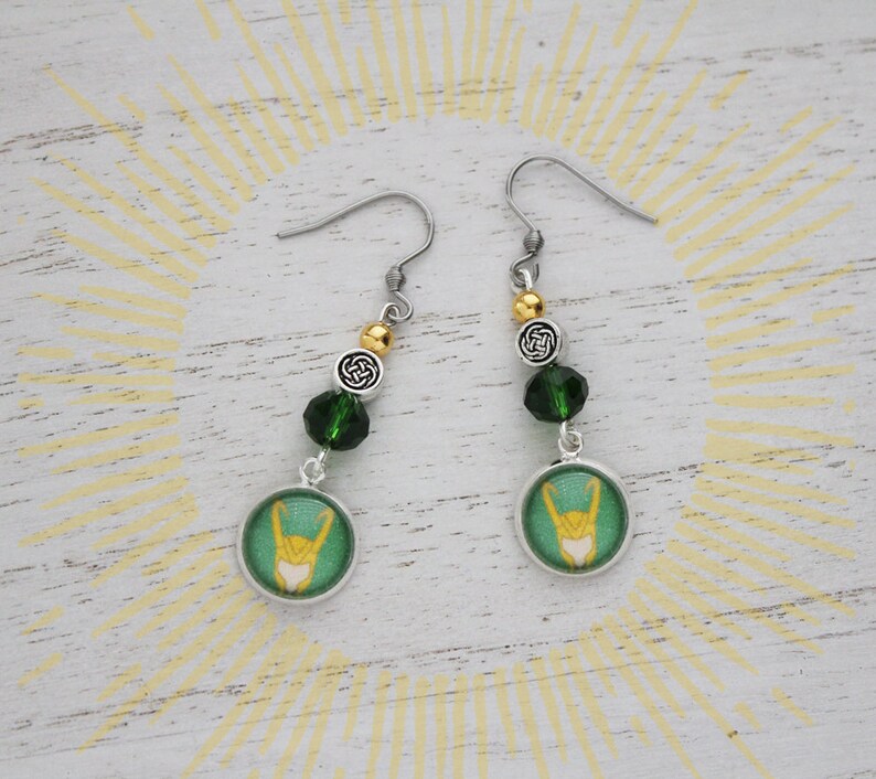 以下が含まれることがあります： 緑と金のアクセントが施されたシルバーカラーのドロップピアス。各イヤリングには、金のデザイン、緑色のビーズ、ケルトノットチャームが付いた円形の緑色のチャームが付いています。スタイリッシュなアクセサリー。
