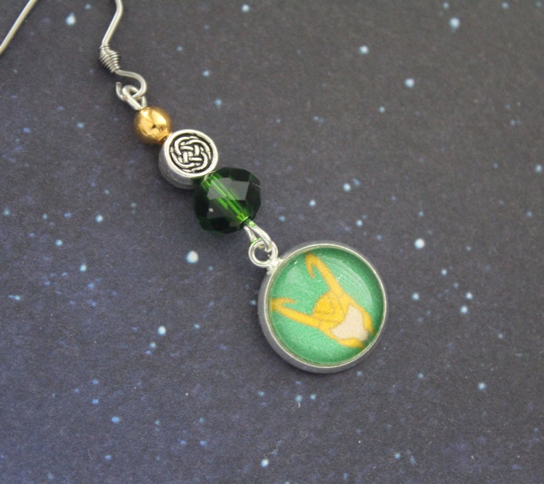 以下が含まれることがあります： 緑と金のデザインのシルバーカラーのドロップイヤリング。イヤリングは、黄色のフィギュア、緑と金のビーズ、ケルトノットチャームを備えた緑と金の円形チャームが特徴です。