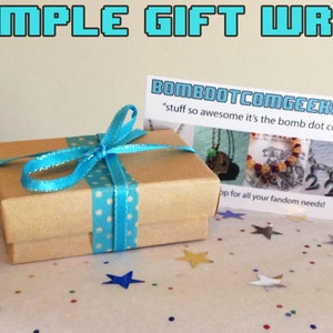 K&ouml;nnte beinhalten: Eine kleine, braune Geschenkbox, die mit einem t&uuml;rkisfarbenen Band mit wei&szlig;en Punkten gebunden ist. Die Box befindet sich auf einer Oberfl&auml;che, die mit sternf&ouml;rmigem Konfetti &uuml;bers&auml;t ist. Eine Karte mit dem Text "BOMBOOTCOMGEEKERY" ist sichtbar.