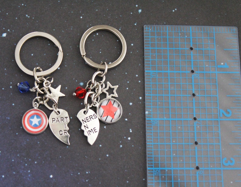 K&ouml;nnte beinhalten: Zwei silberne Schl&uuml;sselanh&auml;nger mit Anh&auml;ngern. Einer hat einen Captain America Schild-Anh&auml;nger, einen Stern-Anh&auml;nger und einen Herz-Anh&auml;nger mit der Aufschrift "PART I CR". Der andere hat einen Stern-Anh&auml;nger, einen Herz-Anh&auml;nger mit der Aufschrift "NERS IN ME" und einen Anh&auml;nger mit einem roten Stern.