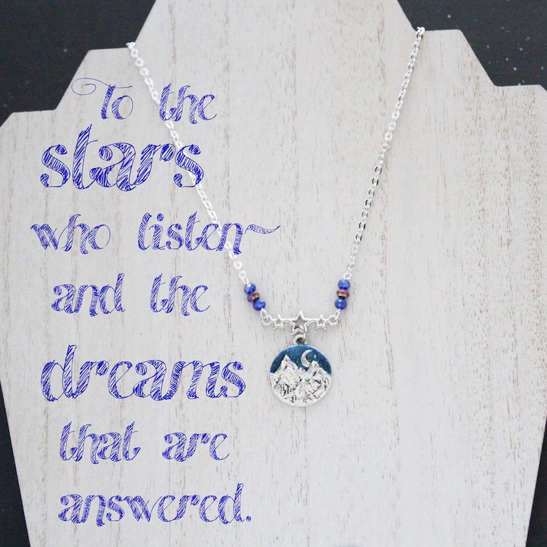 May include: A silver necklace with a blue and silver mountain scene pendant. The necklace has blue beads and a star charm. The necklace is displayed on a white wooden surface with the text "To the stars who listen and the dreams that are answered."