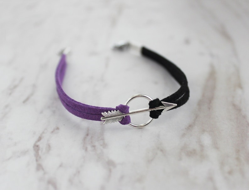 May include: A purple and black suede bracelet with a silver arrow charm and a circle.