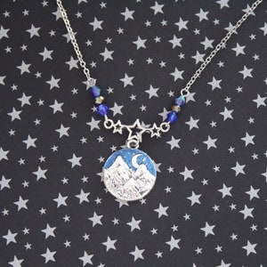 May include: A silver necklace with a blue and white enamel pendant featuring a mountain range and a crescent moon. The necklace is adorned with blue crystal beads and silver star charms.