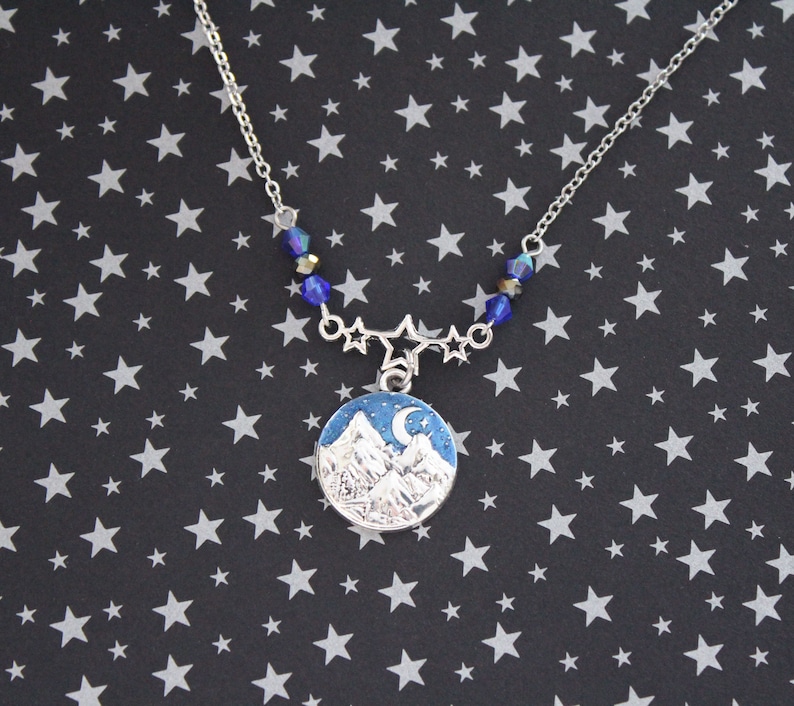 May include: A silver necklace with a blue and white enamel pendant featuring a mountain range and a crescent moon. The necklace is adorned with blue crystal beads and silver star charms.