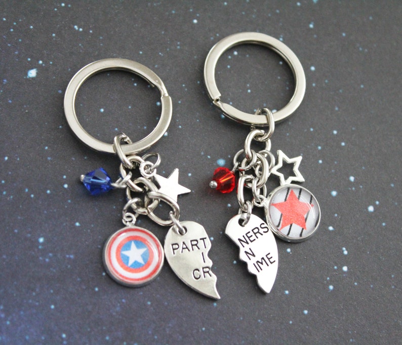 K&ouml;nnte beinhalten: Zwei passende Schl&uuml;sselanh&auml;nger mit silbernen Ringen und Anh&auml;ngern. Einer hat einen Captain America Schild-Anh&auml;nger, eine blaue Kristallperle und einen Stern-Anh&auml;nger. Der andere hat einen roten Stern-Anh&auml;nger, eine rote Kristallperle und einen Stern-Anh&auml;nger. Die Anh&auml;nger lesen "PART I CR" und "NERS N IME".