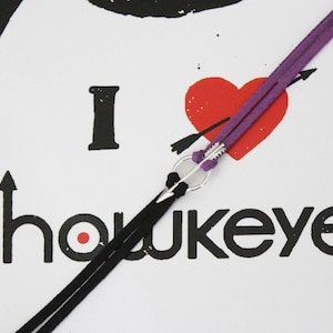 May include: A purple and black leather cord bracelet with a silver arrow charm. The bracelet is tied around a white card with the text "I love Hawkeye" and a red heart with an arrow through it.