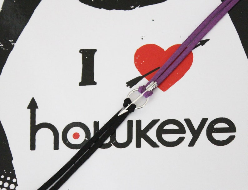 May include: A purple and black leather cord bracelet with a silver arrow charm. The bracelet is tied around a white card with the text "I love Hawkeye" and a red heart with an arrow through it.