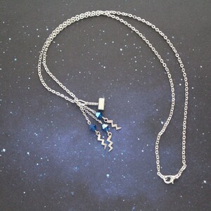 May include: A silver chain necklace with a silver charm of a hammer and blue crystal accents. The necklace is on a dark blue background with a starry pattern.