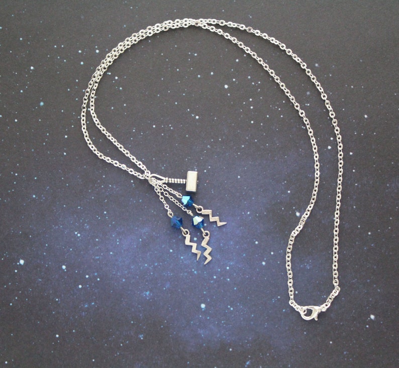 May include: A silver chain necklace with a silver charm of a hammer and blue crystal accents. The necklace is on a dark blue background with a starry pattern.