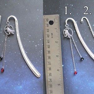 Puede incluir: Dos dise&ntilde;os de marcap&aacute;ginas de plata con un dije de p&aacute;jaro y cuentas colgantes. El marcap&aacute;ginas de la izquierda tiene una cuenta negra y una cuenta roja. El marcap&aacute;ginas de la derecha tiene una cuenta negra y una cuenta roja. Los marcap&aacute;ginas se miden con una regla.