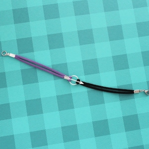 May include: A purple and black leather bracelet with a silver arrow charm. The bracelet is on a turquoise and white checkered background.