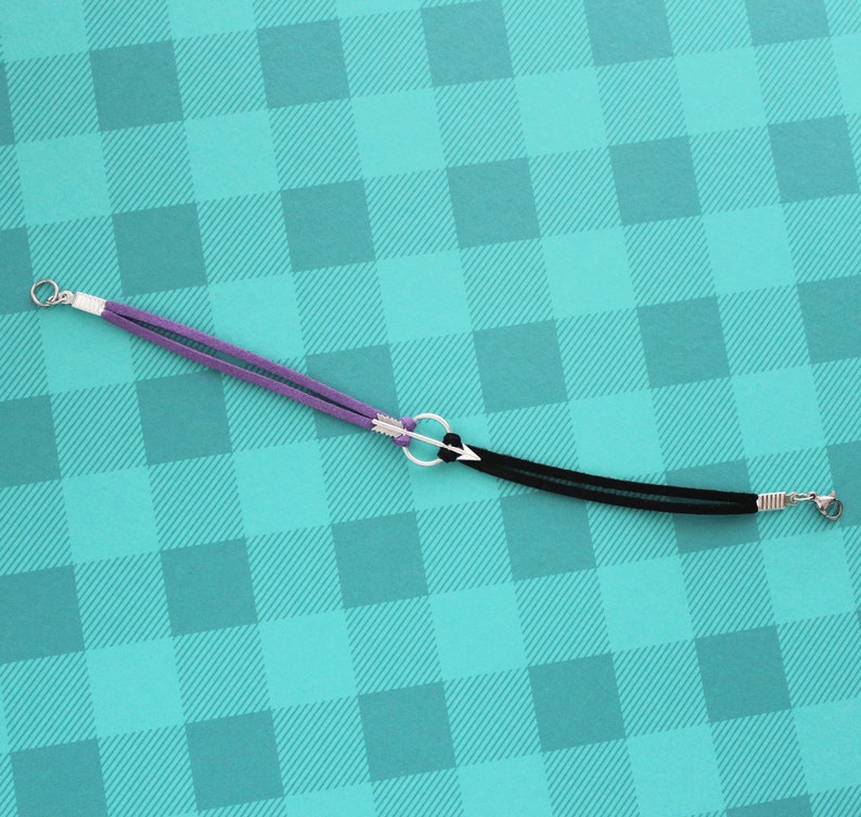 May include: A purple and black leather bracelet with a silver arrow charm. The bracelet is on a turquoise and white checkered background.