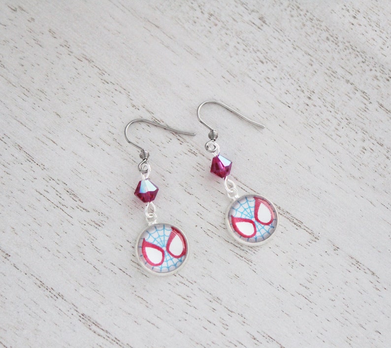 Puede incluir: Un par de pendientes colgantes plateados. Cada pendiente presenta un dise&ntilde;o de telara&ntilde;a roja y blanca en un c&iacute;rculo, acentuado por una peque&ntilde;a cuenta de cristal rosa facetada. Los pendientes se muestran sobre una superficie de madera clara.
