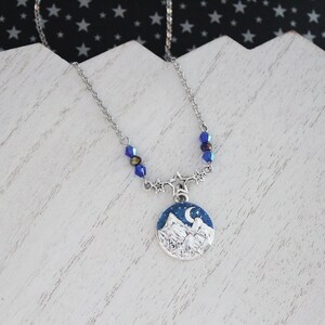 May include: A silver necklace with a blue and white mountain scene pendant. The pendant features a crescent moon and stars. The necklace is adorned with blue crystal beads.