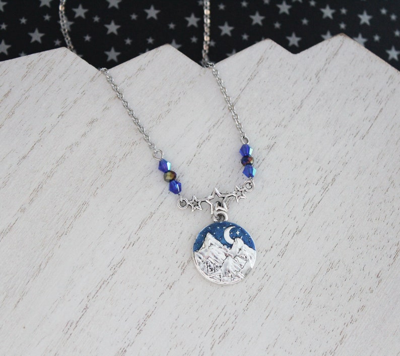 May include: A silver necklace with a blue and white mountain scene pendant. The pendant features a crescent moon and stars. The necklace is adorned with blue crystal beads.