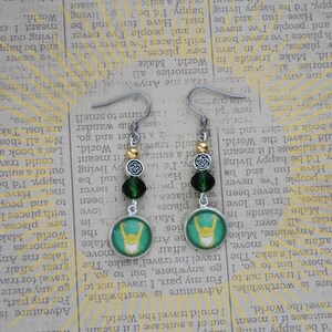 以下が含まれることがあります： 緑と白の円形デザインに様式化されたシンボルが特徴の、シルバーカラーのぶら下がりピアス。ピアスには濃い緑色のビーズと金色のビーズが付いています。ユニークなアクセサリー。