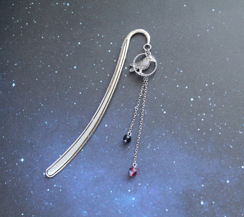 Puede incluir: Marcap&aacute;ginas de plata con un colgante de un p&aacute;jaro con cuentas de cristal negras y rojas.