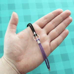May include: A black and purple suede cord bracelet with a silver arrow charm. The arrow charm has a small circle at the top and a feather detail at the bottom.