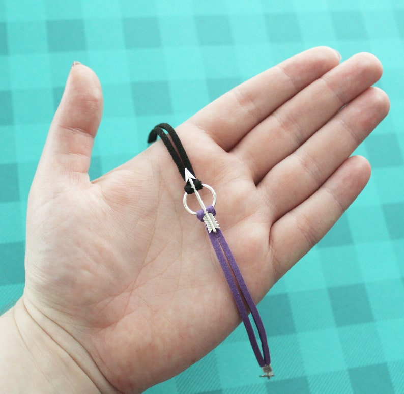 May include: A black and purple suede cord bracelet with a silver arrow charm. The arrow charm has a small circle at the top and a feather detail at the bottom.