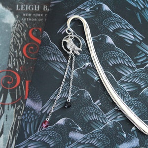 Puede incluir: Marcap&aacute;ginas de plata con un dise&ntilde;o de cuervo y cuentas de cristal rojo y negro colgantes. El marcap&aacute;ginas est&aacute; sobre un fondo de un patr&oacute;n negro y plateado de cuervos.