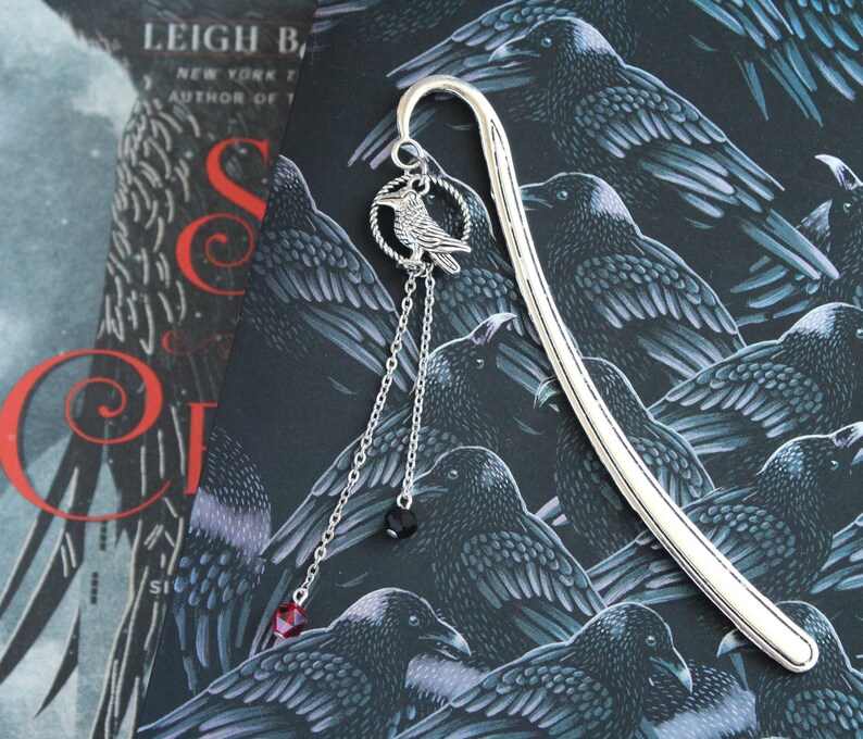 Puede incluir: Marcap&aacute;ginas de plata con un dise&ntilde;o de cuervo y cuentas de cristal rojo y negro colgantes. El marcap&aacute;ginas est&aacute; sobre un fondo de un patr&oacute;n negro y plateado de cuervos.