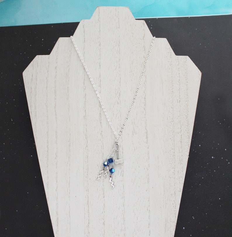 May include: A silver chain necklace with a silver hammer charm and blue crystal beads. The necklace is displayed on a white wooden stand.