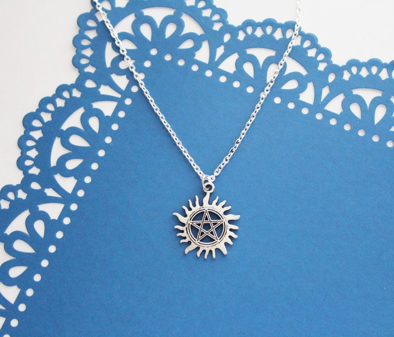 K&ouml;nnte beinhalten: Eine silberne Halskette mit einem Pentagramm-Anh&auml;nger in einem Sonnensymbol. Die Kette liegt auf einem blauen Hintergrund mit einem Spitzenmuster.