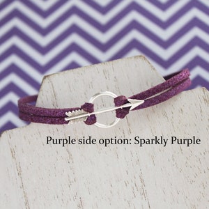 May include: A purple suede bracelet with a silver arrow charm and a circle. The bracelet is on a white display stand. Purple side option: Sparkly Purple.