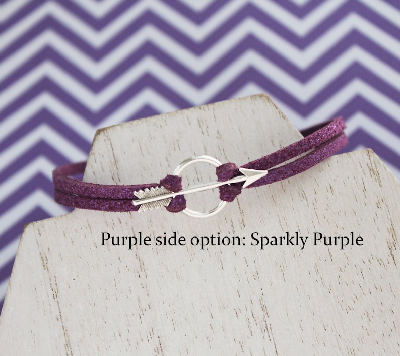 May include: A purple suede bracelet with a silver arrow charm and a circle. The bracelet is on a white display stand. Purple side option: Sparkly Purple.