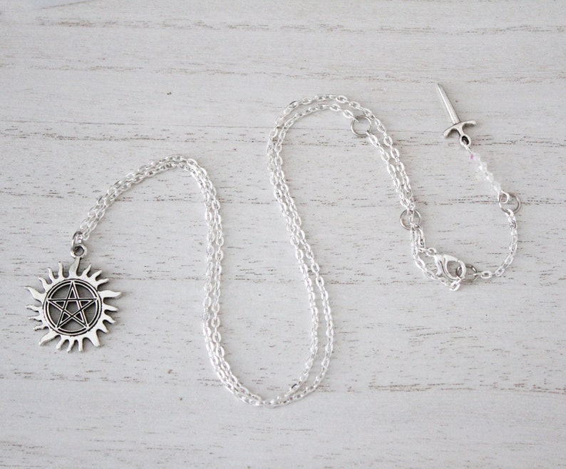 K&ouml;nnte beinhalten: Eine silberne Halskette mit einem Pentagramm-Anh&auml;nger und einem passenden silbernen Schwertanh&auml;nger. Die Halskette liegt auf einer wei&szlig;en Holzoberfl&auml;che.