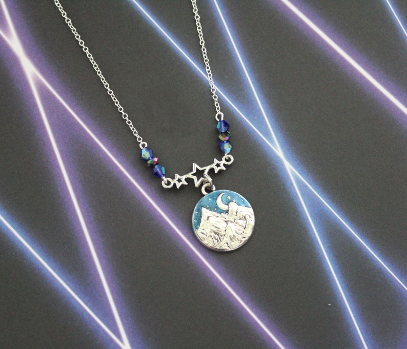 May include: A silver necklace with a blue and white enamel pendant featuring a crescent moon and mountains. The necklace is adorned with blue crystal beads and silver star charms.