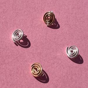 May include: Four silver and gold spiral earrings on a pink background. The earrings are all different sizes and shapes.