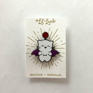 May include: A white enamel pin featuring a cartoon white cat with purple wings and a red heart on its head. The pin is on a white card with gold lettering that reads "The Lil' Lambs" and the text "@the.lil.lamb thelillamb.com".