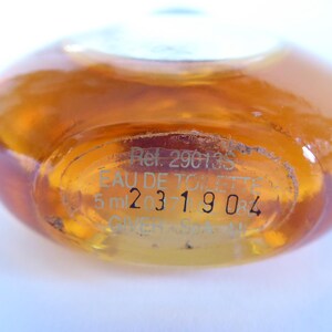 K&ouml;nnte beinhalten: Eine kleine, runde, bernsteinfarbene Glasflasche mit Fl&uuml;ssigkeit. Der Boden ist mit Text graviert, darunter "Ref. 29013S", "EAU DE TOILETTE", "5 ml" und die Zahlenfolge "231904". Die Flasche hat eine glatte, gew&ouml;lbte Oberfl&auml;che aus Klarglas.