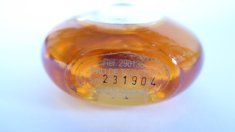 K&ouml;nnte beinhalten: Eine kleine, runde, bernsteinfarbene Glasflasche mit Fl&uuml;ssigkeit. Der Boden ist mit Text graviert, darunter "Ref. 29013S", "EAU DE TOILETTE", "5 ml" und die Zahlenfolge "231904". Die Flasche hat eine glatte, gew&ouml;lbte Oberfl&auml;che aus Klarglas.