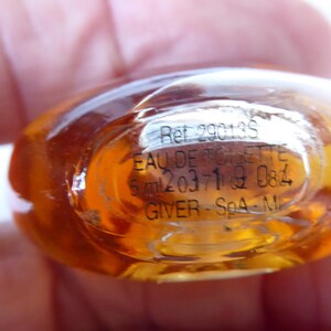 K&ouml;nnte beinhalten: Ein kleiner, ovaler Glasparf&uuml;mflakon mit einer bernsteinfarbenen Fl&uuml;ssigkeit. Der Boden zeigt aufgedruckten Text, darunter Ref 29013S, EAU DE TOILETTE, 5 ml, 203719 084 und GIVER - SpA - MI, der durch den klaren Glasboden sichtbar ist.