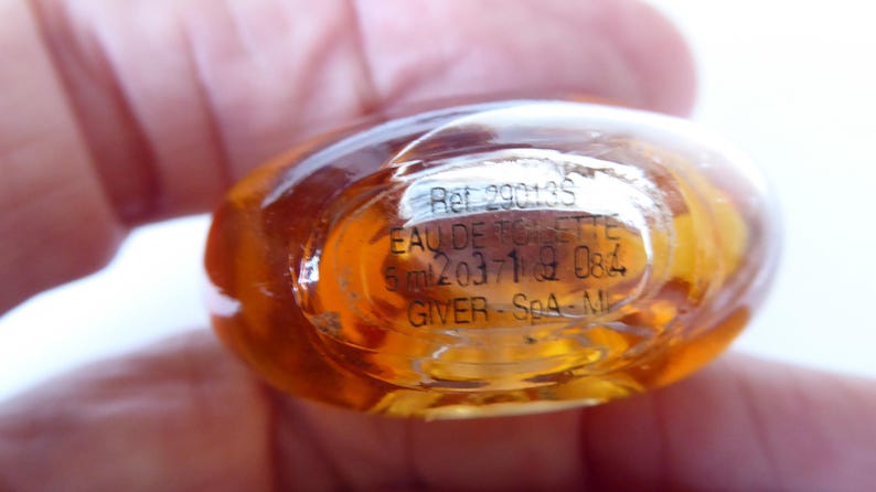 K&ouml;nnte beinhalten: Ein kleiner, ovaler Glasparf&uuml;mflakon mit einer bernsteinfarbenen Fl&uuml;ssigkeit. Der Boden zeigt aufgedruckten Text, darunter Ref 29013S, EAU DE TOILETTE, 5 ml, 203719 084 und GIVER - SpA - MI, der durch den klaren Glasboden sichtbar ist.