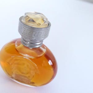 K&ouml;nnte beinhalten: Ein kleiner, ovaler Parf&uuml;mflakon aus Glas, gef&uuml;llt mit bernsteinfarbener Fl&uuml;ssigkeit. Der Flakon hat einen silberfarbenen Verschluss mit einem griechischen M&auml;andermuster am Rand und einen klaren, facettierten, geometrischen Aufsatz.