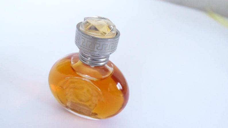 K&ouml;nnte beinhalten: Ein kleiner, ovaler Parf&uuml;mflakon aus Glas, gef&uuml;llt mit bernsteinfarbener Fl&uuml;ssigkeit. Der Flakon hat einen silberfarbenen Verschluss mit einem griechischen M&auml;andermuster am Rand und einen klaren, facettierten, geometrischen Aufsatz.