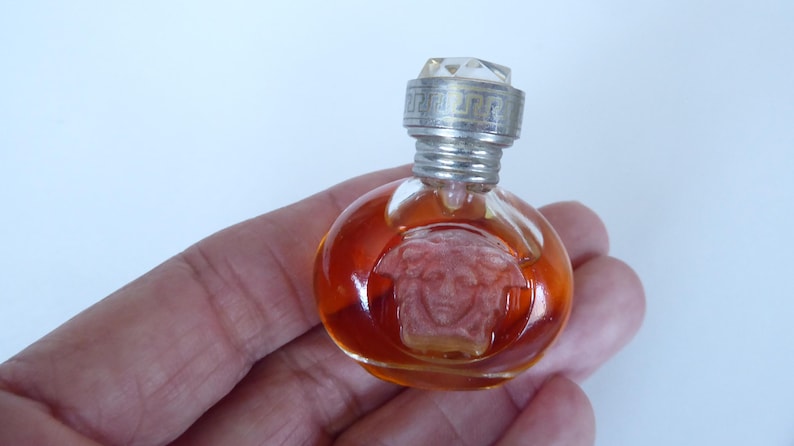 K&ouml;nnte beinhalten: Ein kleiner, runder Parf&uuml;mflakon aus klarem Glas, gef&uuml;llt mit bernsteinfarbener Fl&uuml;ssigkeit. Die Vorderseite des Flakons zeigt ein eingepr&auml;gtes Medusenhaupt-Relief. Er hat einen silberfarbenen Metalldeckel mit M&auml;ander-Muster und Kristallaufsatz.