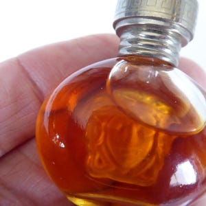 K&ouml;nnte beinhalten: Ein kleines, rundes Parf&uuml;mfl&auml;schchen aus bernsteinfarbenem Glas mit einem metallischen Schraubverschluss. Die Flasche zeigt ein geformtes Reliefdesign eines Gesichts auf der Vorderseite, enth&auml;lt eine goldbraune Fl&uuml;ssigkeit und hat einen gerippten Hals.
