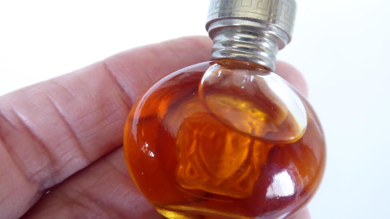 K&ouml;nnte beinhalten: Ein kleines, rundes Parf&uuml;mfl&auml;schchen aus bernsteinfarbenem Glas mit einem metallischen Schraubverschluss. Die Flasche zeigt ein geformtes Reliefdesign eines Gesichts auf der Vorderseite, enth&auml;lt eine goldbraune Fl&uuml;ssigkeit und hat einen gerippten Hals.