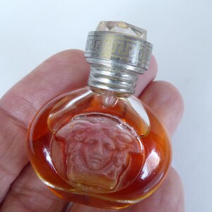 K&ouml;nnte beinhalten: Ein Miniatur-Parf&uuml;mflakon aus Glas mit bernsteinfarbener Fl&uuml;ssigkeit. Der Flakon zeigt ein gepr&auml;gtes Medusenhaupt-Emblem auf der Vorderseite und einen silberfarbenen Metallverschluss mit griechischem M&auml;ander-Muster und klarem Kristallaufsatz.