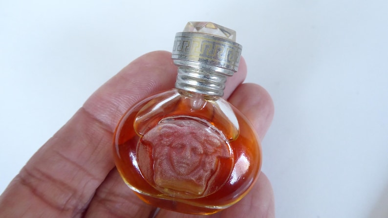 K&ouml;nnte beinhalten: Ein Miniatur-Parf&uuml;mflakon aus Glas mit bernsteinfarbener Fl&uuml;ssigkeit. Der Flakon zeigt ein gepr&auml;gtes Medusenhaupt-Emblem auf der Vorderseite und einen silberfarbenen Metallverschluss mit griechischem M&auml;ander-Muster und klarem Kristallaufsatz.
