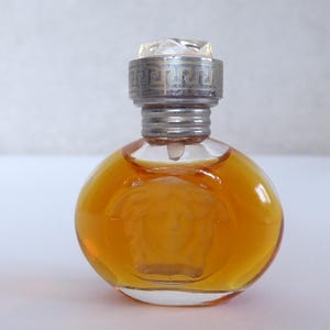 K&ouml;nnte beinhalten: Ein kleiner, runder Parf&uuml;mflakon aus klarem Glas, gef&uuml;llt mit bernsteinfarbener Fl&uuml;ssigkeit. Der Flakon zeigt ein gepr&auml;gtes Medusenhaupt auf der Vorderseite und einen silberfarbenen Metalldeckel mit M&auml;ander-Muster und facettiertem Kristall.