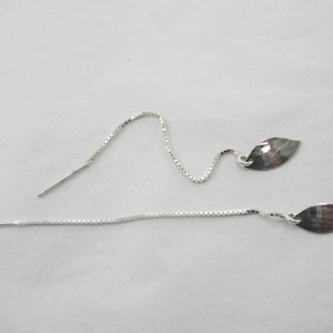 May include: A pair of silver threader earrings. Each earring features a delicate chain that ends in a leaf-shaped charm. The earrings are displayed on a white surface.