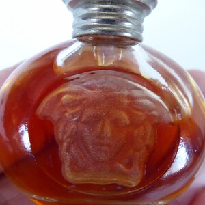 K&ouml;nnte beinhalten: Ein kleiner, runder Parf&uuml;mflakon aus bernsteinfarbenem Glas mit einem erhabenen, mattierten Medusenhaupt-Emblem auf der Vorderseite. Der Flakon hat einen metallisch silberfarbenen Schraubverschluss und enth&auml;lt eine durch das Glas sichtbare orangefarbene Fl&uuml;ssigkeit.