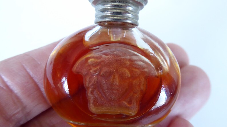 K&ouml;nnte beinhalten: Ein kleiner, runder Parf&uuml;mflakon aus bernsteinfarbenem Glas mit einem erhabenen, mattierten Medusenhaupt-Emblem auf der Vorderseite. Der Flakon hat einen metallisch silberfarbenen Schraubverschluss und enth&auml;lt eine durch das Glas sichtbare orangefarbene Fl&uuml;ssigkeit.